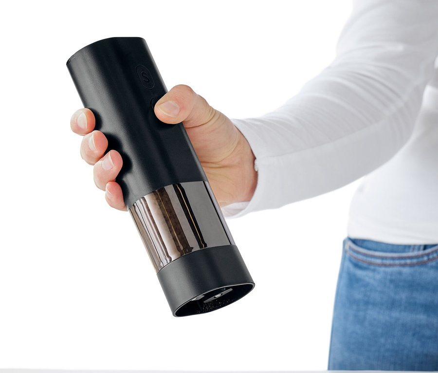 Eine Hand hält eine schwarze Gewürzmühle über einen weißen Tisch, aus der Gewürze rieseln.