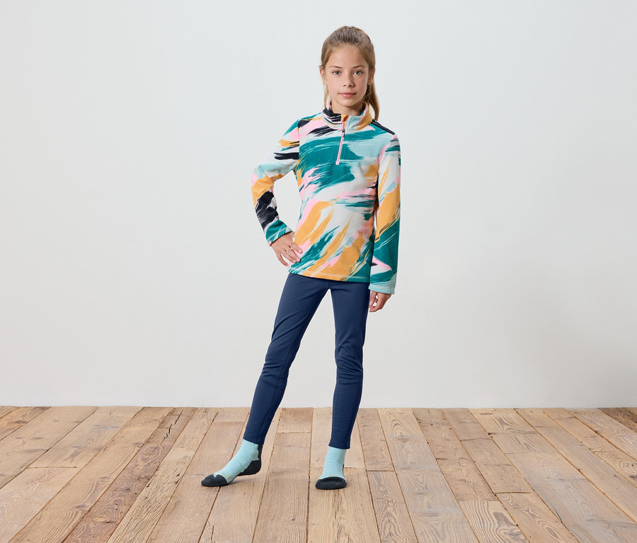Ein Mädchen in einem bunten Langarmshirt, dunkelblauen Hosen und Socken steht auf einem Holzboden.