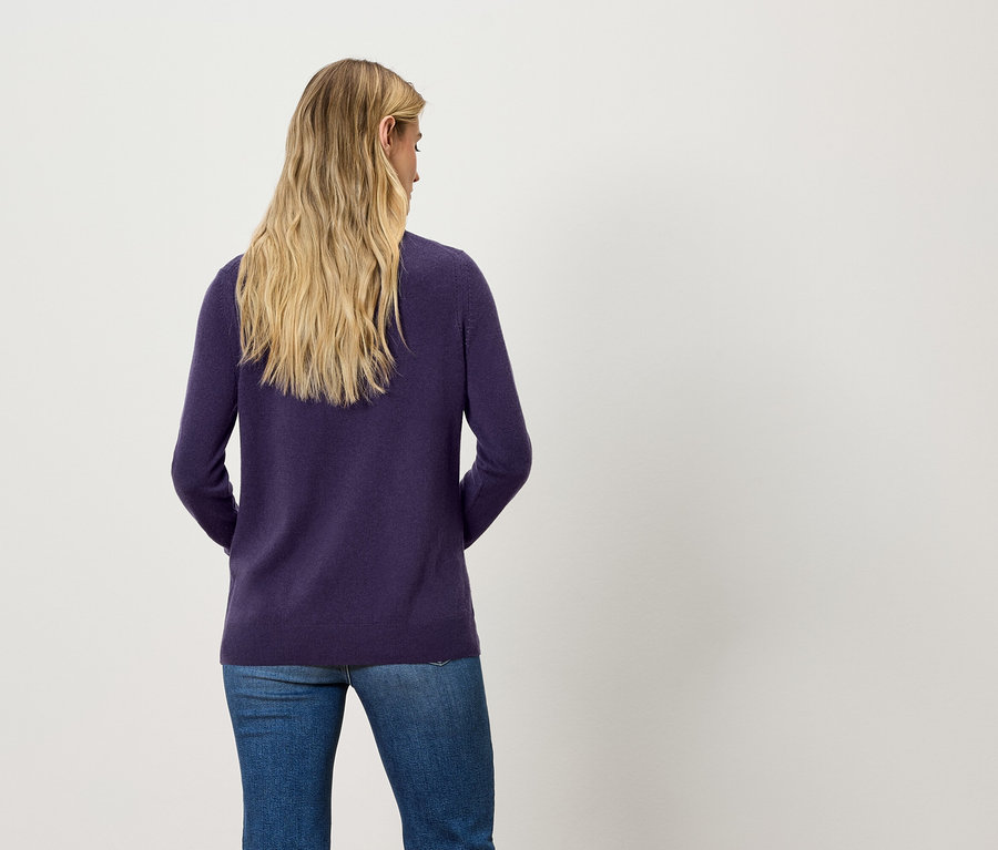 Rückansicht einer blonden Frau in einem dunkelvioletten Pullover und blauen Jeans.