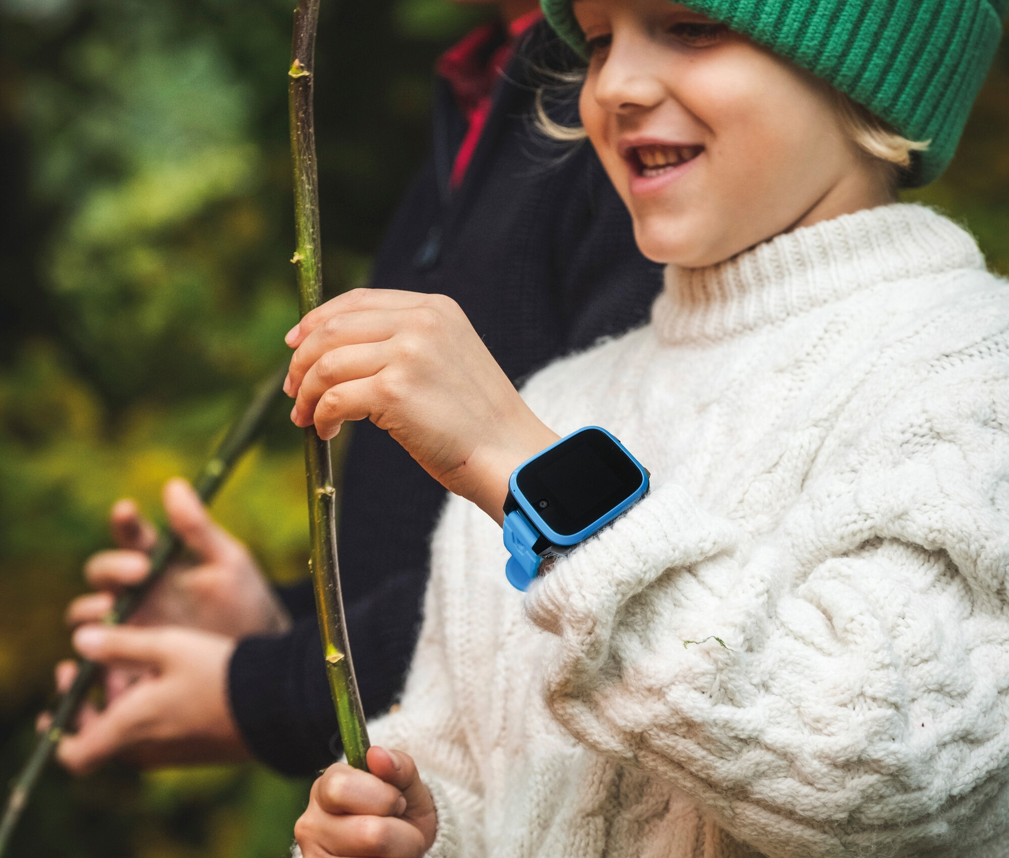Junge mit blauem Kids Watch Xplora XGO3 und grüner Mütze hält einen Stock in der Hand.