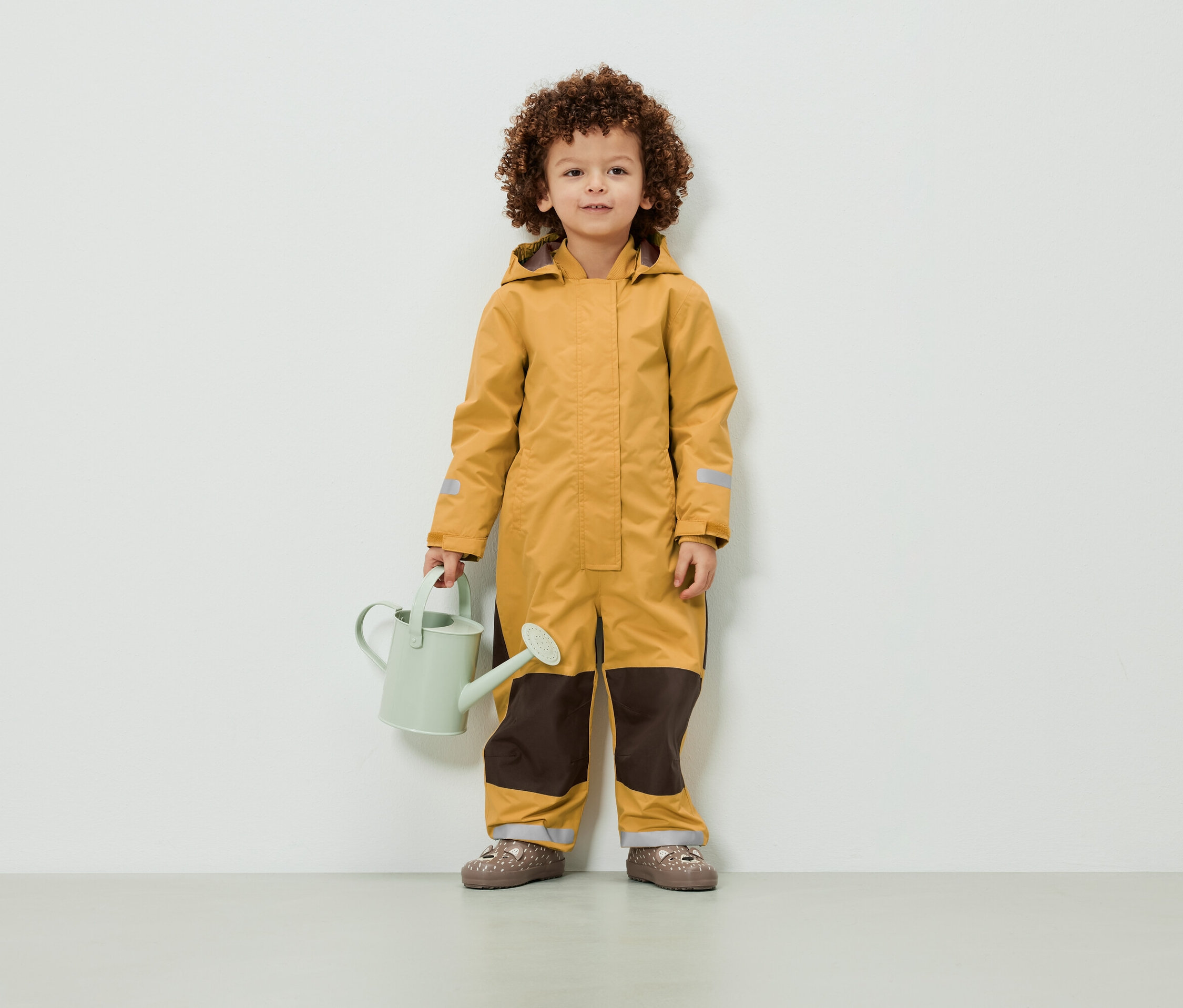 Ein Kind mit lockigem Haar steht in einem gelben Overall und hält eine hellgrüne Gießkanne.