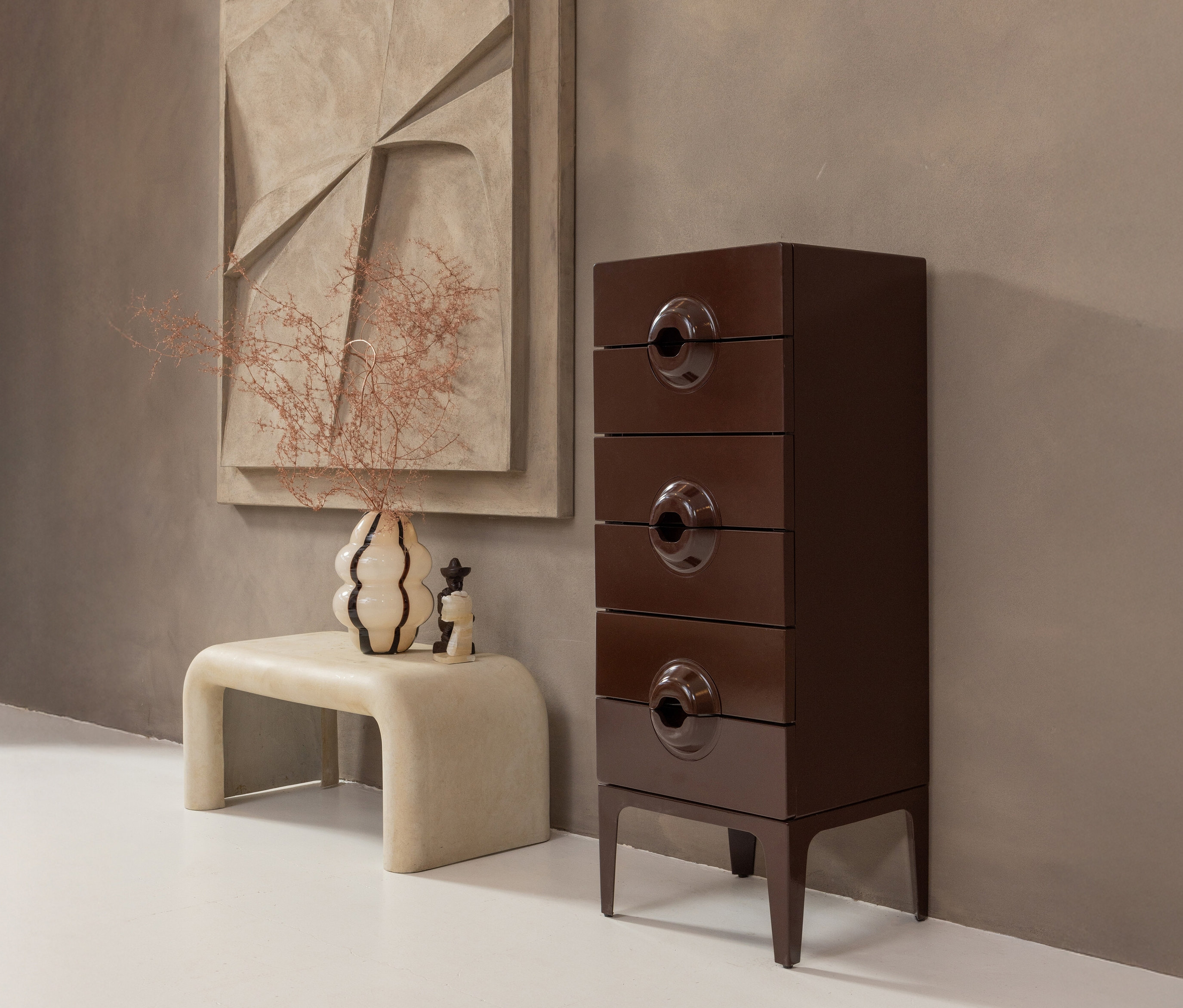 Eine rote Woood Kommode »Merge« steht neben einem beigen Hocker mit einer Vase und Trockenblumen.