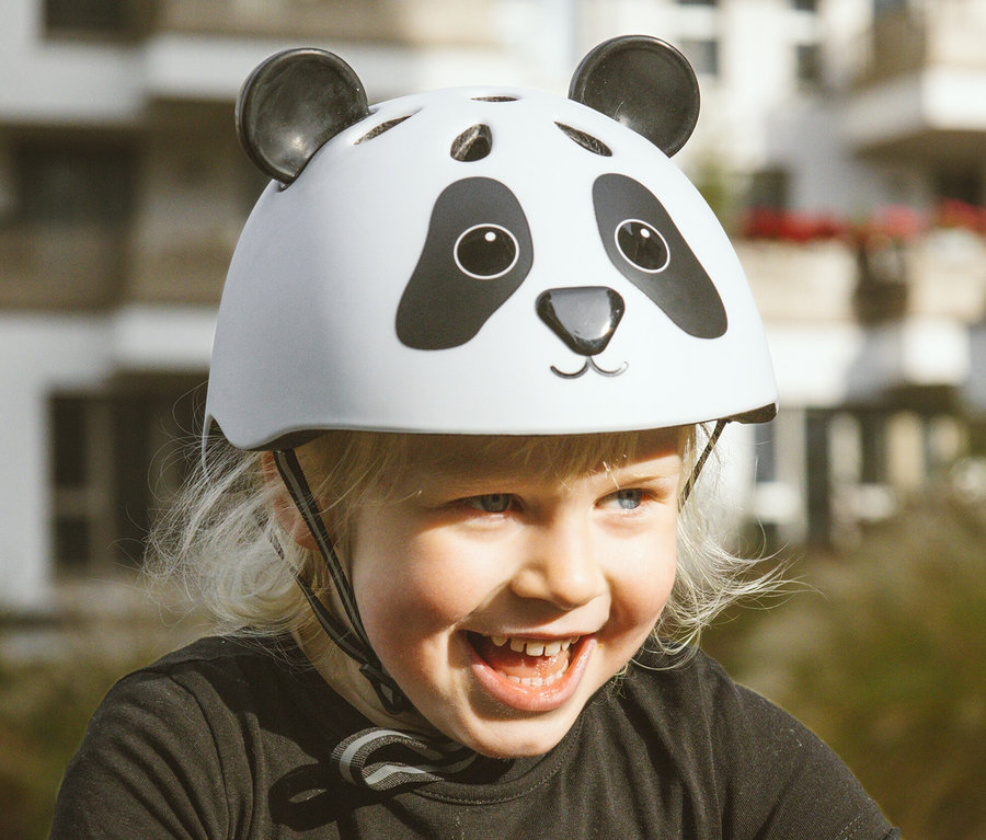 Lächelndes blondes Mädchen trägt ein schwarzes Hemd mit Skelett-Aufdruck und einen HUDORA Kinderhelm »Pandy« mit Panda-Motiv.