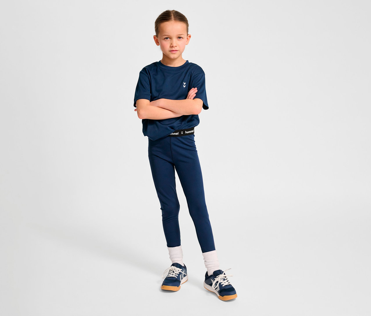 Mädchen posiert mit verschränkten Armen in marineblauem T-Shirt und HUMMEL HMLJR Pulse Mid Waist Tight Leggings.