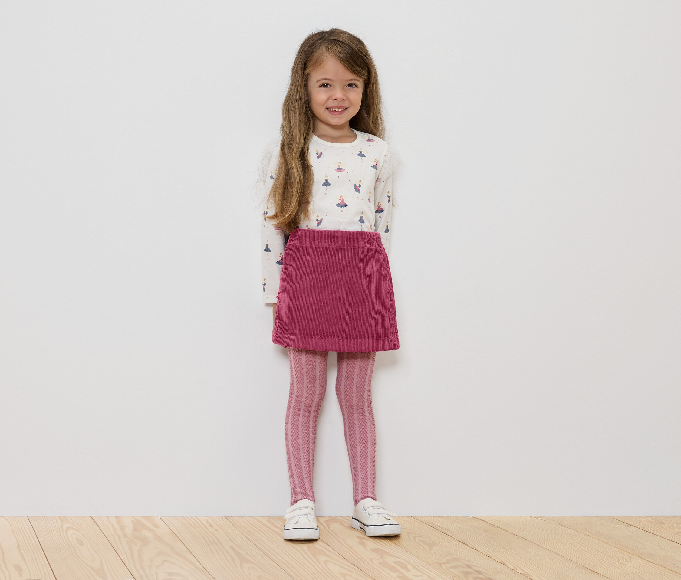 Ein Mädchen steht und trägt ein weißes, langärmliges Hemd mit Ballerinas, einen rosafarbenen Cordrock, rosa Leggings mit Muster und weiße Turnschuhe.