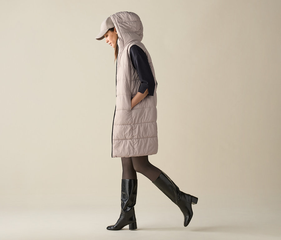 Eine Frau geht und trägt eine gesteppte Weste mit Kapuze, eine Mütze und Kniestiefel.