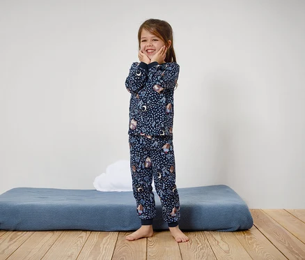 Ein lächelndes Mädchen in einem blauen Pyjama mit Aufdruck steht auf einem Holzboden.