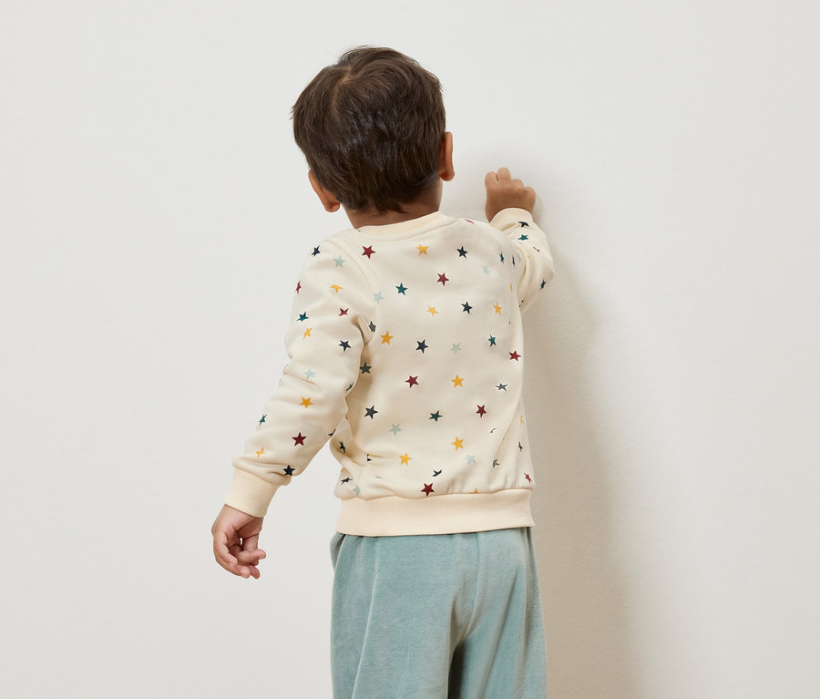 Junge in cremefarbenem Sternenpullover steht an der Wand.