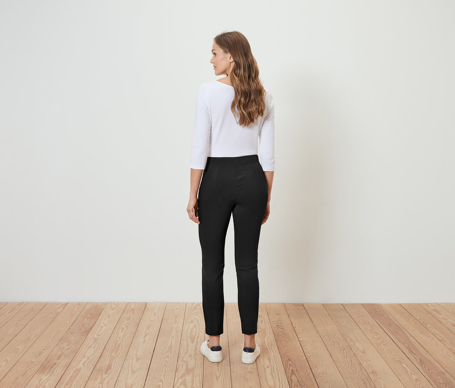 Eine Frau steht mit dem Rücken zur Kamera und trägt eine braune Hose und eine weiße Bluse.