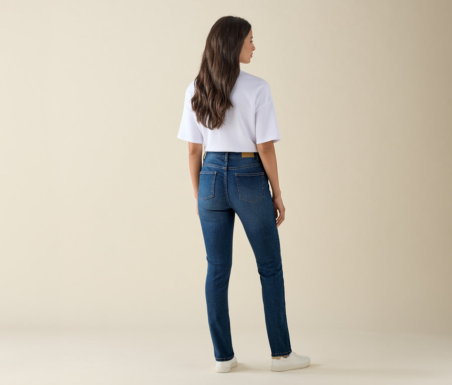 Eine Frau steht mit dem Rücken zur Kamera und trägt eine Slim-Jeans – Fit »Emma«, Mid Blue Denim, ein weißes T-Shirt und Turnschuhe.