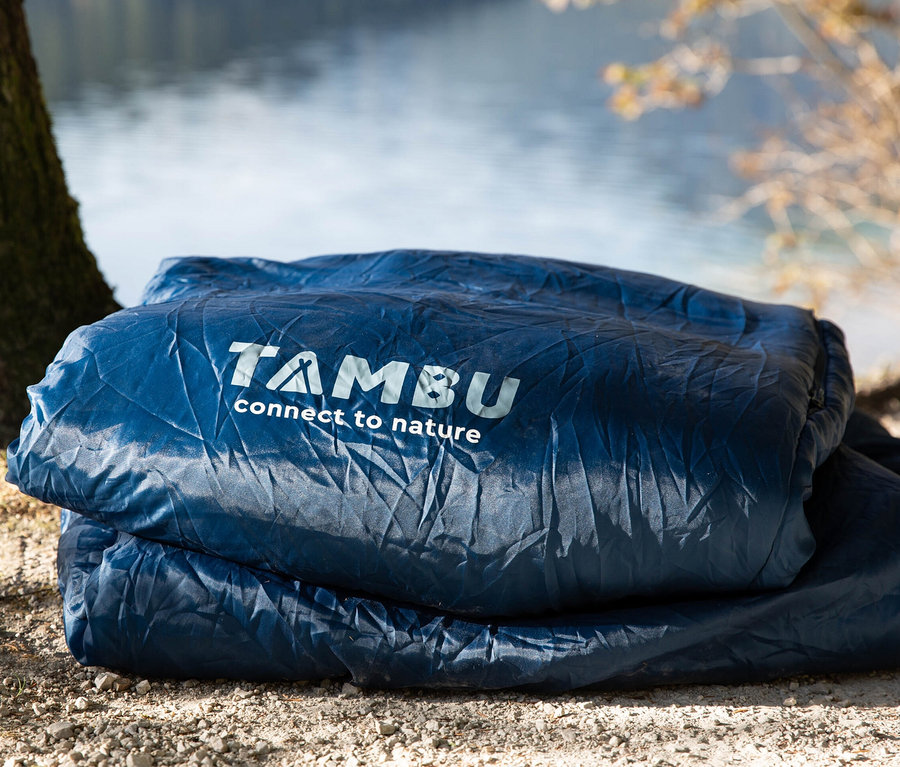 Ein blauer Tambu Mumienschlafsack »TALU 1500« liegt neben einem Baum auf dem Boden.