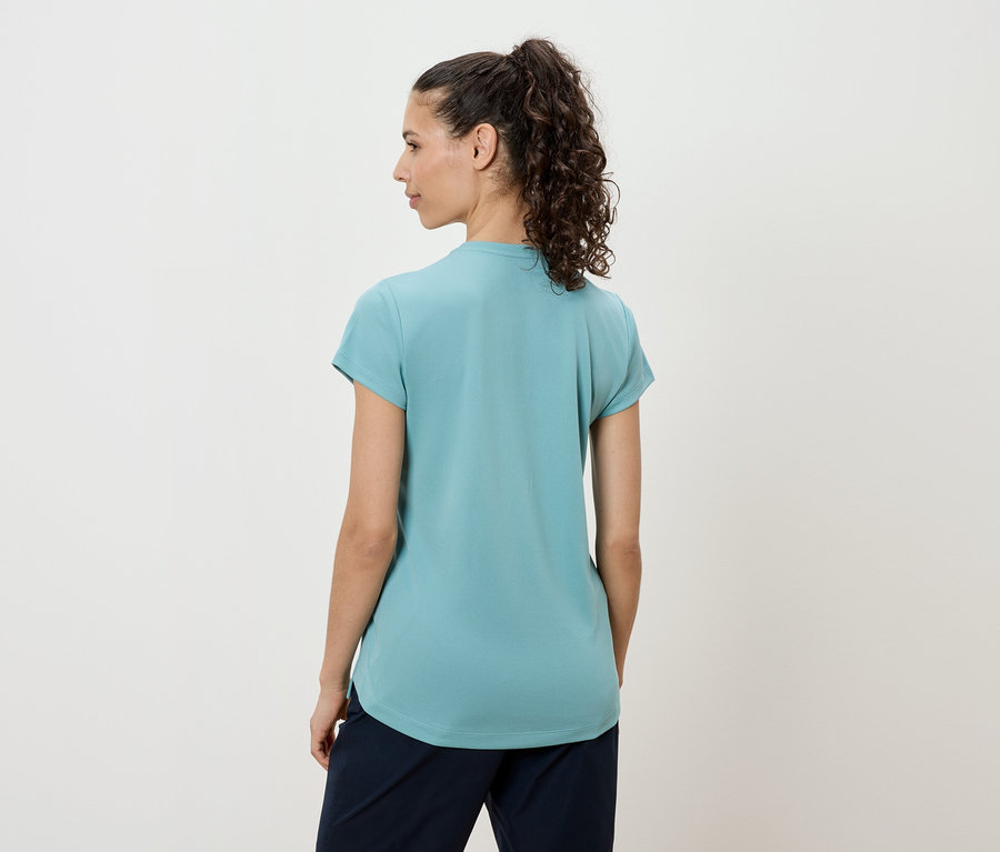 Frau mit dunklen, lockigen Haaren trägt ein türkisfarbenes T-Shirt und eine dunkle Hose, von hinten gesehen.