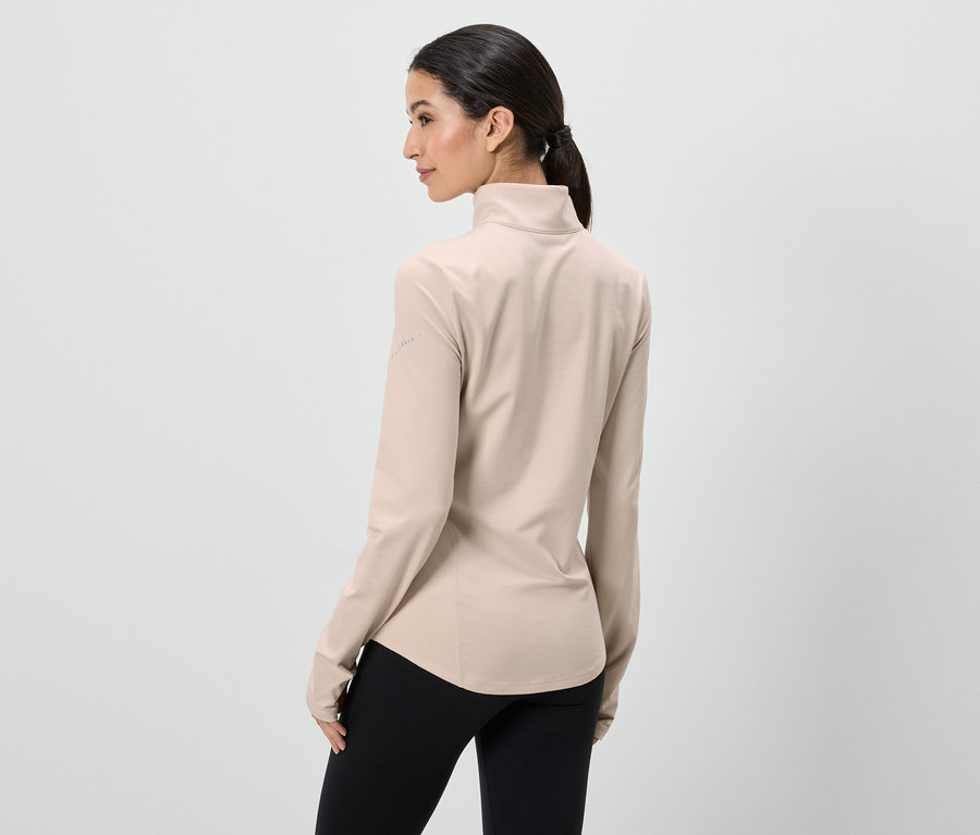 Frau von hinten, trägt beigefarbenes, langärmliges Oberteil und schwarze Leggings vor einem weißen Hintergrund.