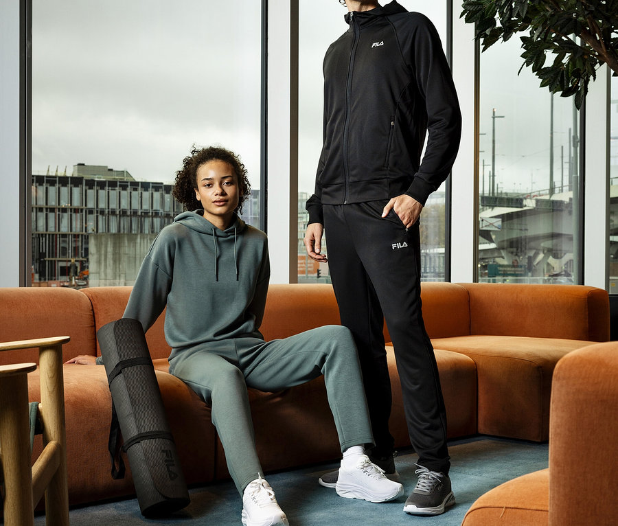 Frau sitzt mit FILA Damen-Hoodie aus Modalmix und Yogamatte auf Couch. Mann steht daneben in Trainingsjacke und -hose.