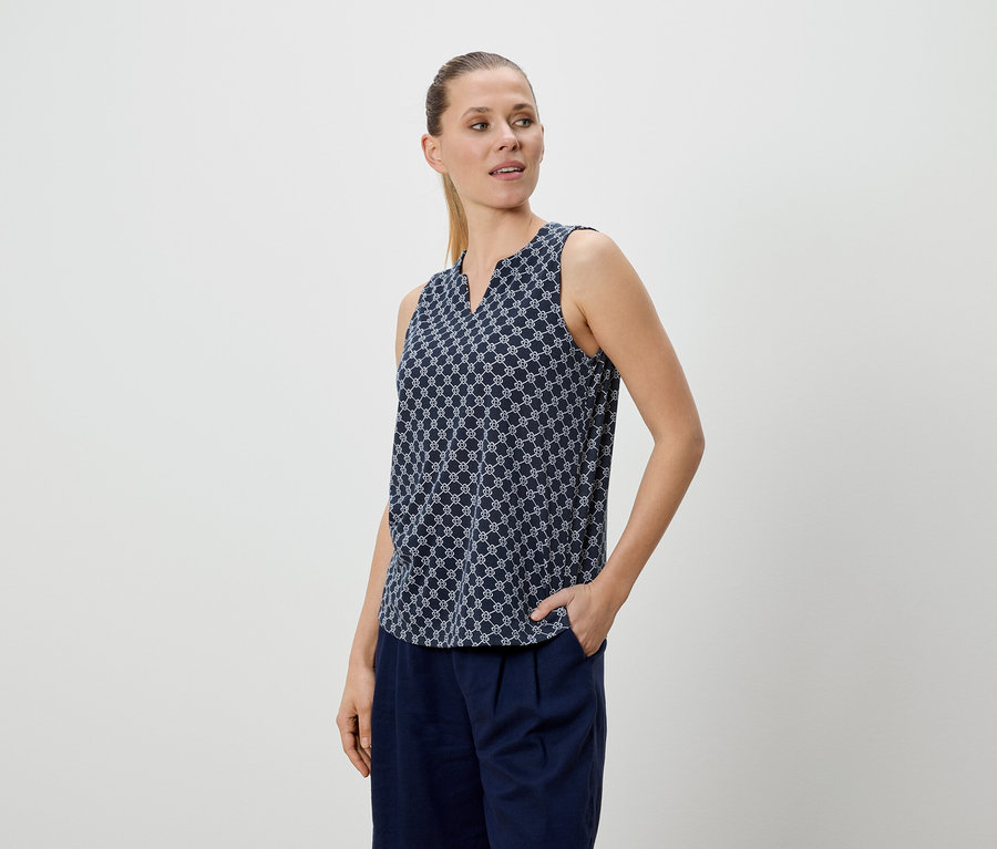 Frau mit blau gemusterter Bluse und blauer Hose, eine Hand in der Hosentasche.
