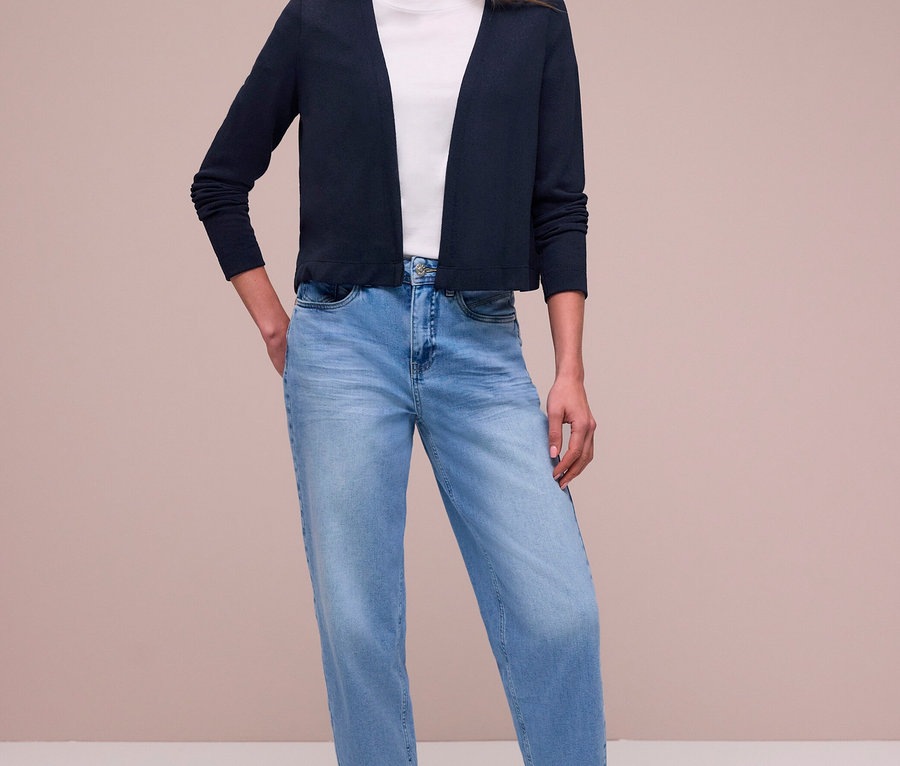 Frau steht mit Händen in den Taschen, trägt einen dunkelblauen Street One Cardigan über einem weißen T-Shirt, hellblaue Jeans und silberne Sneaker.