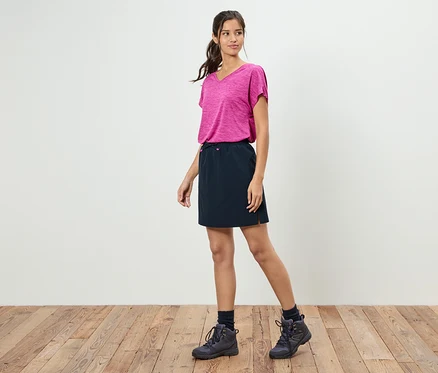 Eine Frau steht vor einer weißen Wand und trägt ein pinkfarbenes T-Shirt, einen schwarzen Rock und Wanderschuhe.