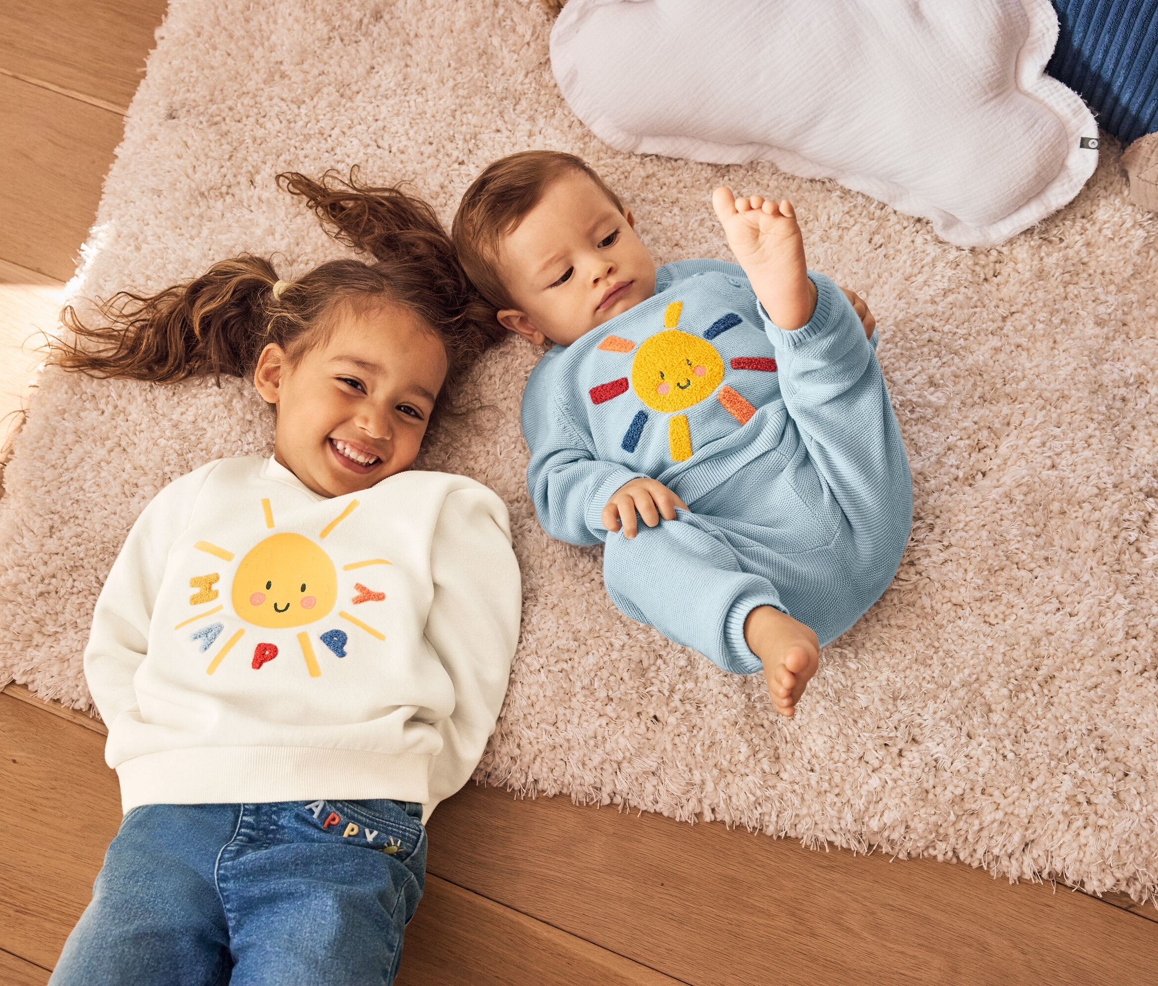 Ein Mädchen mit Zöpfen und ein Baby liegen auf einem Teppich. Das Mädchen trägt ein cremefarbenes Sweatshirt mit einer gelben Sonne. Das Baby trägt einen blauen Strickanzug mit einer Sonne.