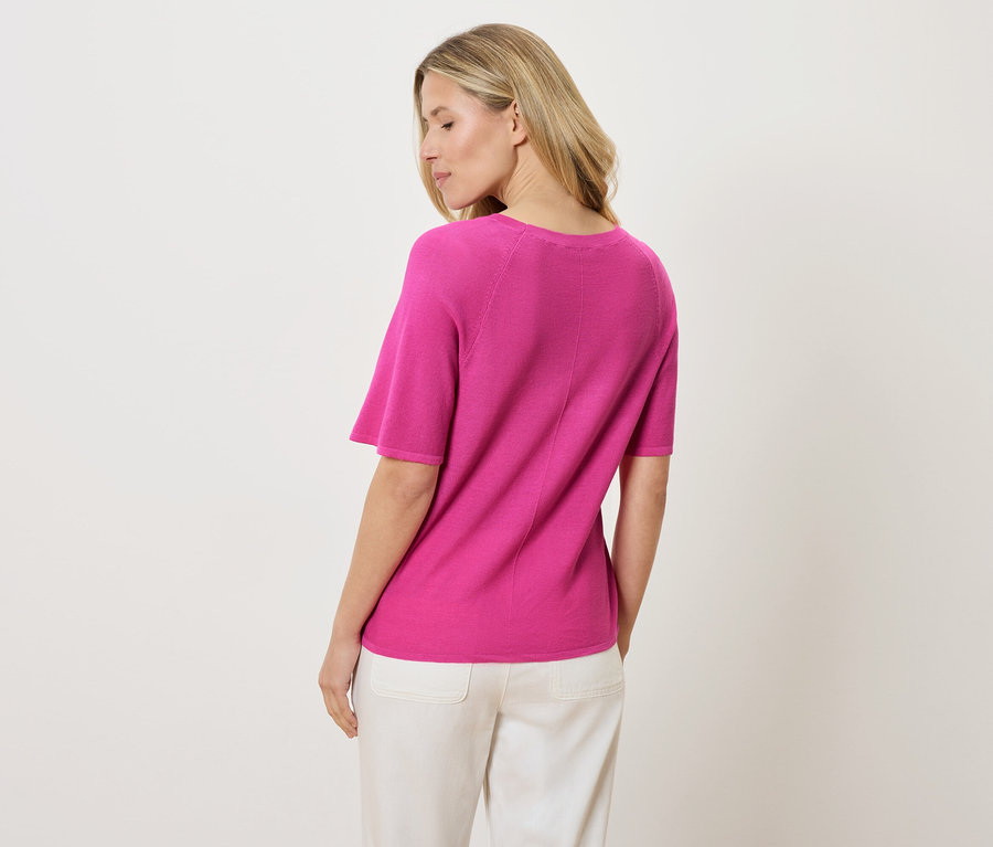 Rückansicht einer Frau mit blonden Haaren, die ein pinkes Kurzarm-Shirt und weiße Hosen trägt.