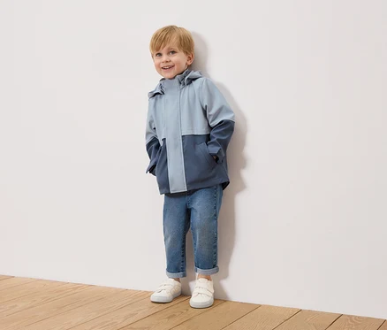 Ein lächelnder Junge mit blonden Haaren steht vor einer weißen Wand und trägt eine zweifarbige, hellblaue und marineblaue Jacke, blaue Jeans und weiße Turnschuhe.