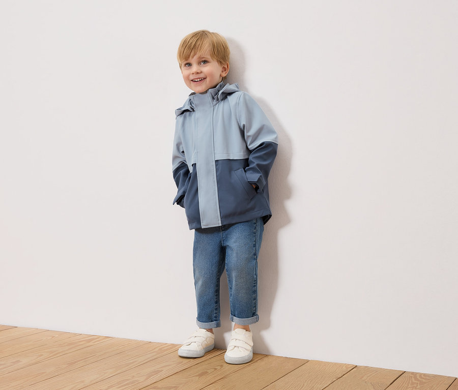 Ein lächelnder Junge mit blonden Haaren steht vor einer weißen Wand und trägt eine zweifarbige, hellblaue und marineblaue Jacke, blaue Jeans und weiße Turnschuhe.