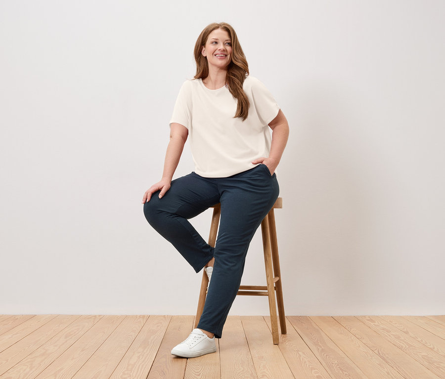 Frau sitzt auf einem Hocker, trägt ein cremefarbenes T-Shirt, blaue Hose und weiße Turnschuhe.