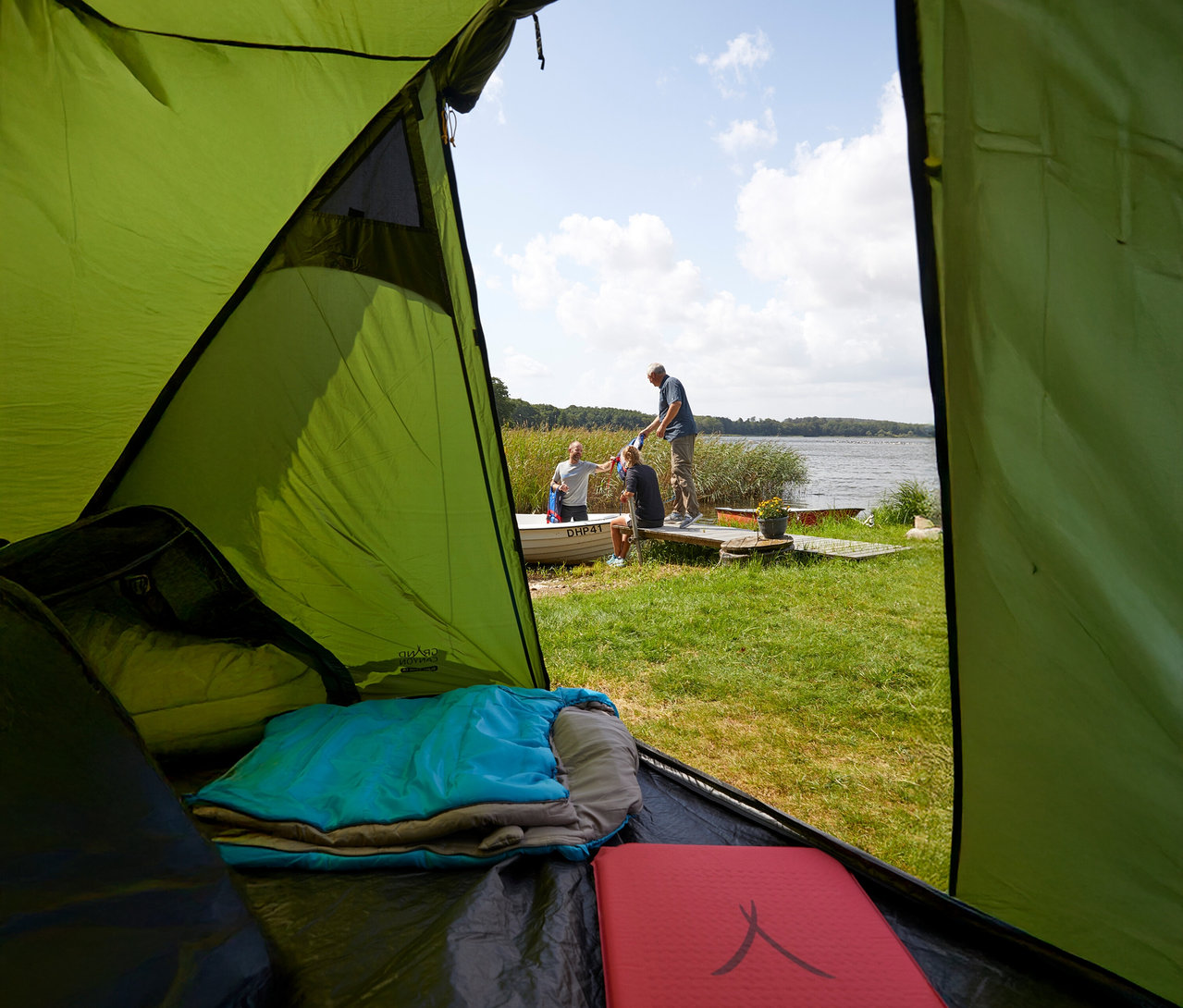 Blick aus einem Zelt auf drei Personen an einem See, mit einer blauen Schlafsackrolle und einer roten Isomatte im Vordergrund.