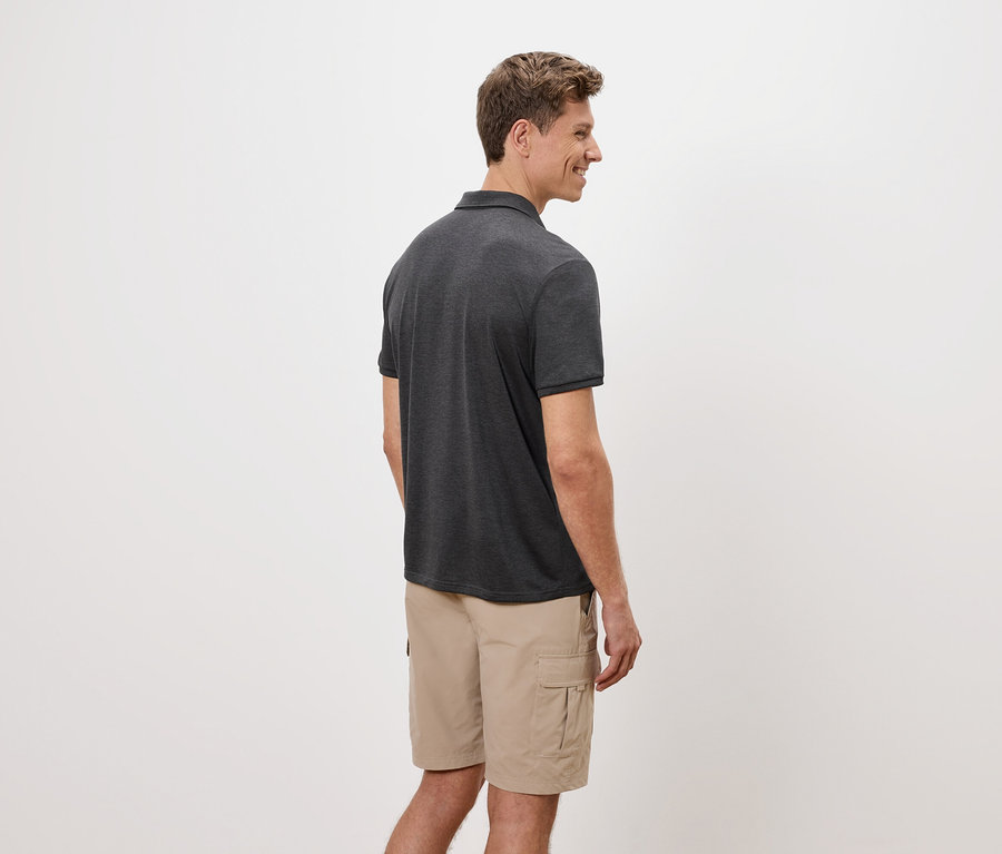 Ein Mann steht seitlich, trägt ein graues Poloshirt und beigefarbene Cargoshorts vor einem weißen Hintergrund.