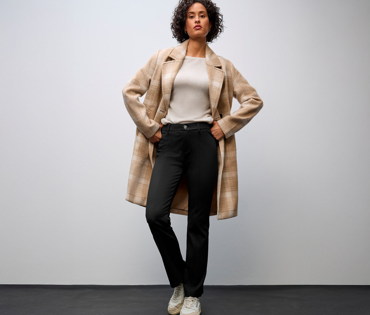 Frau mit beigefarbenem Mantel und Street One Bootcut Hose, die ihre Hände auf die Hüften legt.