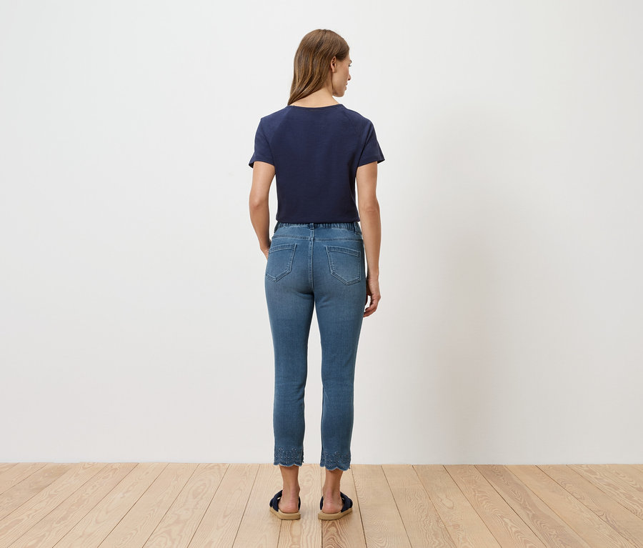 Rückansicht einer Frau mit blauem T-Shirt und Jeans vor einer weißen Wand.