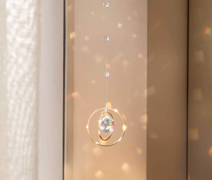 Ein Kristall-Mobile mit goldenen Ringen hängt vor einem Fenster und wirft Lichtflecken an die Wand.