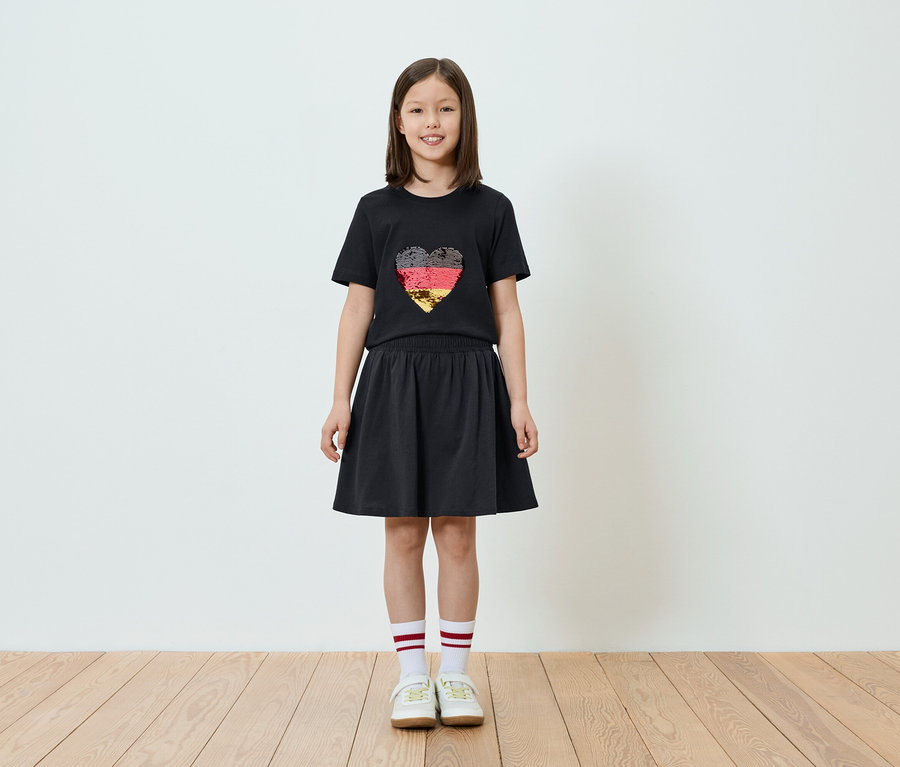 Mädchen in einem schwarzen T-Shirt mit einem Herz in den Farben der deutschen Flagge und einem schwarzen Rock, dazu weiße Socken mit roten Streifen und Turnschuhe.