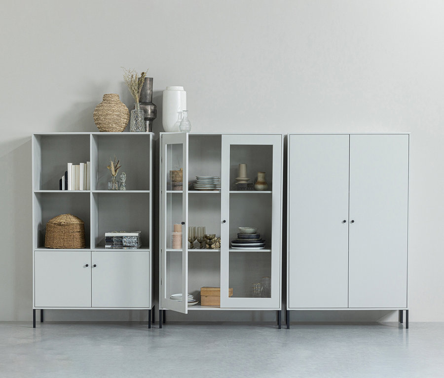 Graue Regale und ein Schrank stehen an einer Wand. Das Regal links hat offene Fächer mit Büchern und Dekorationen. Das mittlere Regal hat eine Glastür, die teilweise geöffnet ist und Geschirr zeigt. Rechts steht ein geschlossener Schrank.