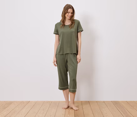 Eine Frau steht barfuß auf einem Holzboden und trägt einen olivgrünen Pyjama mit weißen Akzenten.