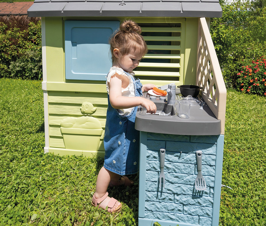 Mädchen spielt am Smoby Life »Pretty« Spielhaus mit Küche im Garten. Sie hantiert mit Spielzeuggeschirr.