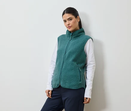 Frau mit Pferdeschwanz, die eine grüne Fleeceweste über einem weißen Langarmshirt und einer marineblauen Hose trägt.