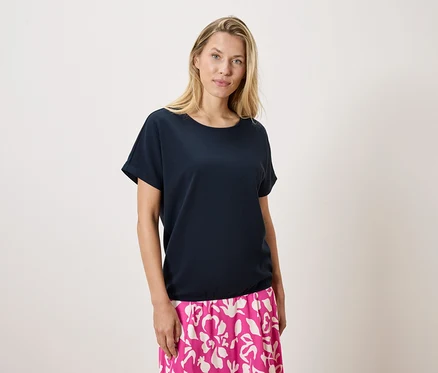 Eine blonde Frau trägt ein marineblaues T-Shirt und einen rosa-weiß gemusterten Rock vor einem weißen Hintergrund.