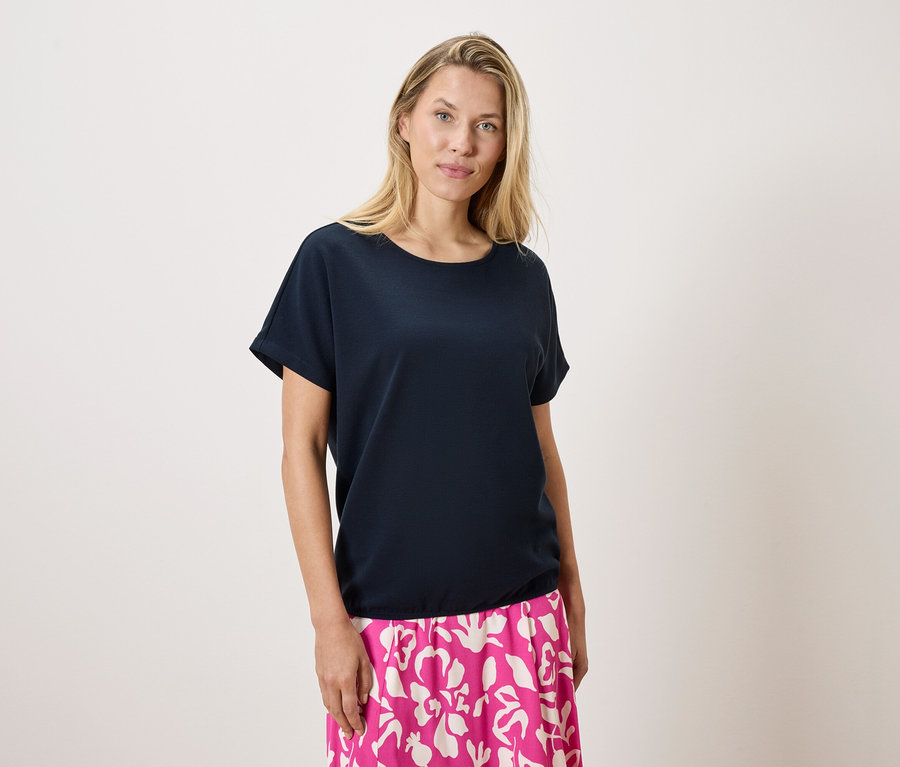 Eine blonde Frau trägt ein marineblaues T-Shirt und einen rosa-weiß gemusterten Rock vor einem weißen Hintergrund.