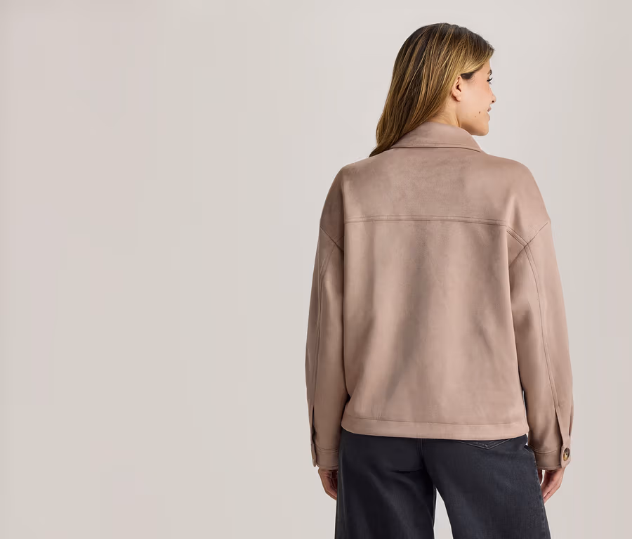 Frau von hinten, trägt eine hellbraune Kragenjacke in Wildleder-Optik und dunkle Hose, blickt nach rechts.