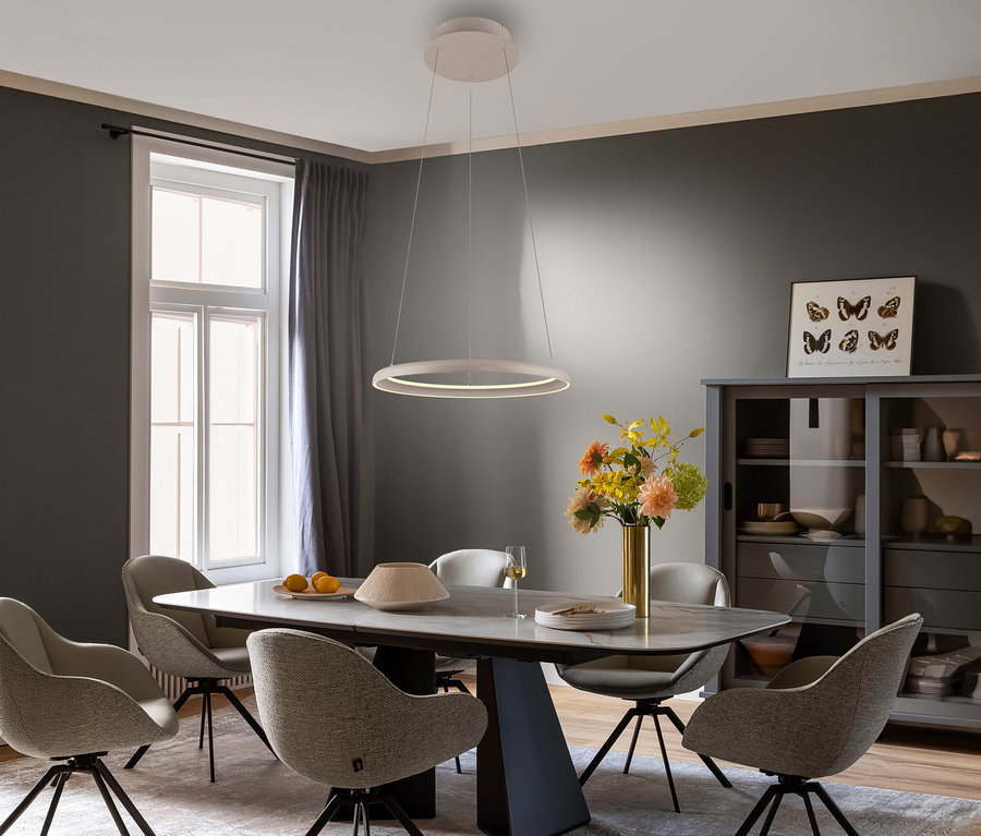 Ein modernes Esszimmer mit einem großen Esstisch und sechs gepolsterten Stühlen. Über dem Tisch hängt eine sandfarbene SCHÖNER WOHNEN-Kollektion Pendelleuchte »Quillo« mit beleuchtetem Innenring. Auf dem Tisch stehen Blumen, Zitronen und Geschirr. Links ist ein Fenster mit grauen Vorhängen, rechts ein grauer Schrank mit Schmetterlingsbildern.