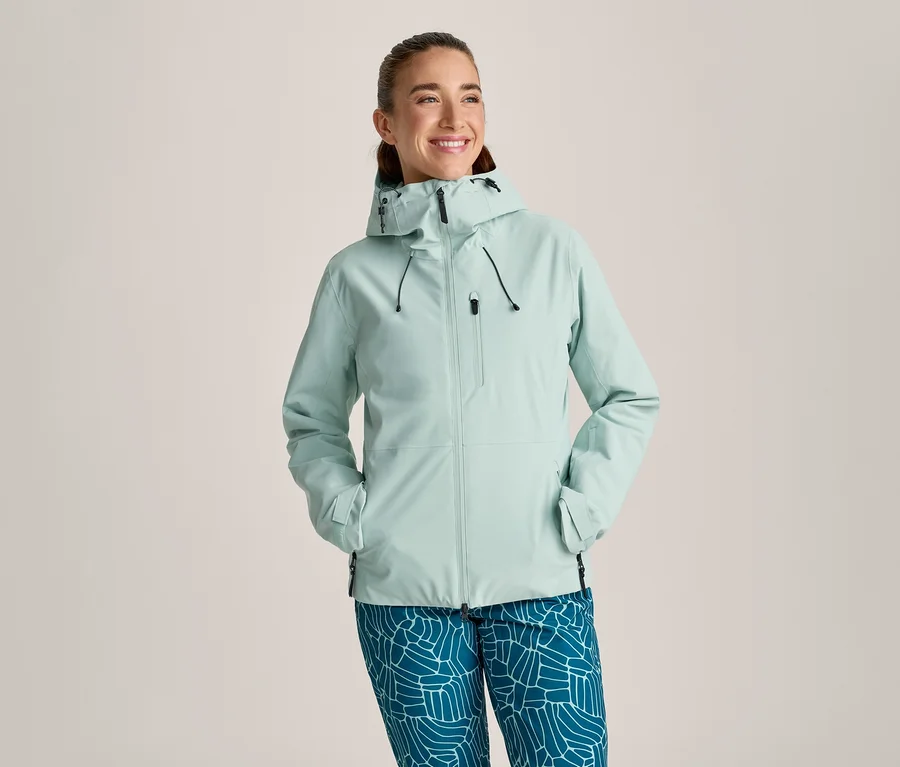 Lächelnde Frau in einer hellen mintgrünen Kapuzenjacke mit schwarzen Reißverschlüssen und Kordelzügen sowie blauen Hosen mit weißem geometrischem Muster. Ihre Hände stecken in den Jackentaschen.