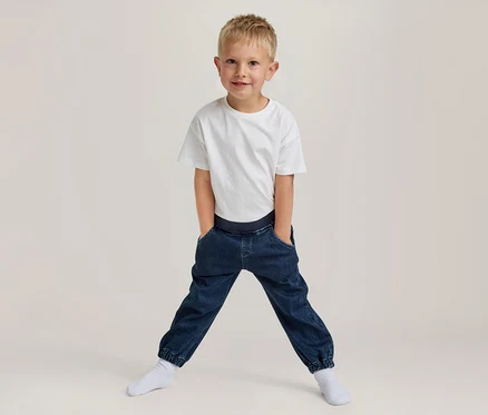 Junger Junge mit blondem Haar, lächelt, trägt ein weißes T-Shirt und dunkelblaue Jogger-Hosen mit den Händen in den Taschen, steht breitbeinig in weißen Socken.