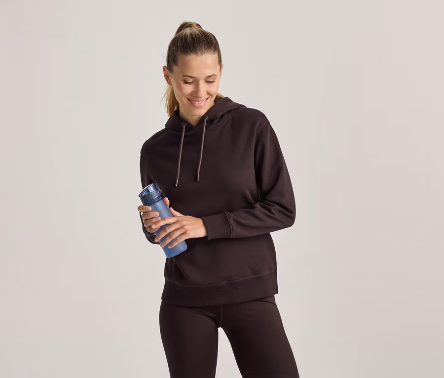 Frau in einem braunen Hoodie und braunen Leggings hält eine blaue Trinkflasche, lächelt und blickt nach unten.