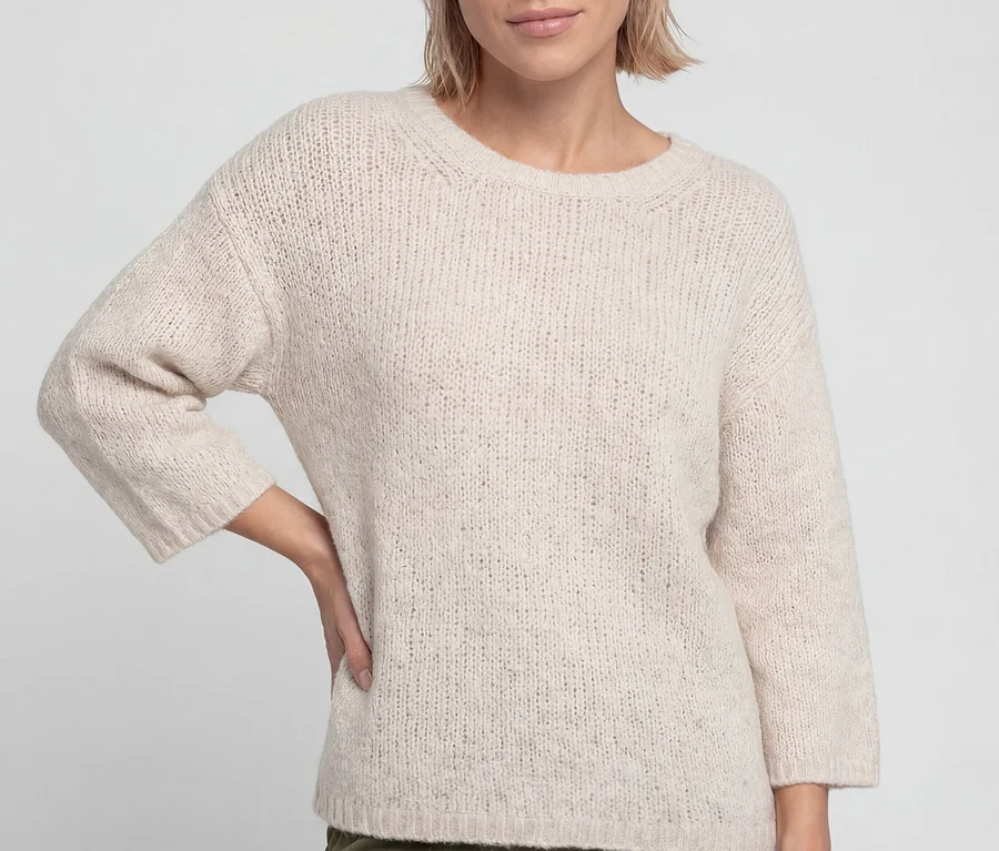 Frau mit kurzem blondem Haar, die einen hellbeigen Olsen 3/4-Pullover und olivgrüne Hosen trägt, steht mit einer Hand in der Hüfte.