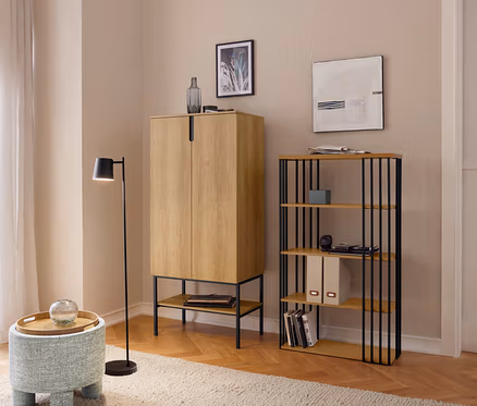 Ein Wohnzimmer mit hellem Holzparkett und beigem Teppich, das einen hellbraunen Schrank, ein Regal mit schwarzen Metallstreben und einen blauen Hocker mit Holztablett zeigt. Eine schwarze Stehlampe steht daneben, und ein Fenster mit Vorhängen ist links sichtbar.