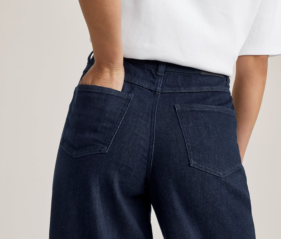 Rückenansicht einer Person in dunkelblauen Jeans und weißem T-Shirt, eine Hand steckt in der Gesäßtasche.