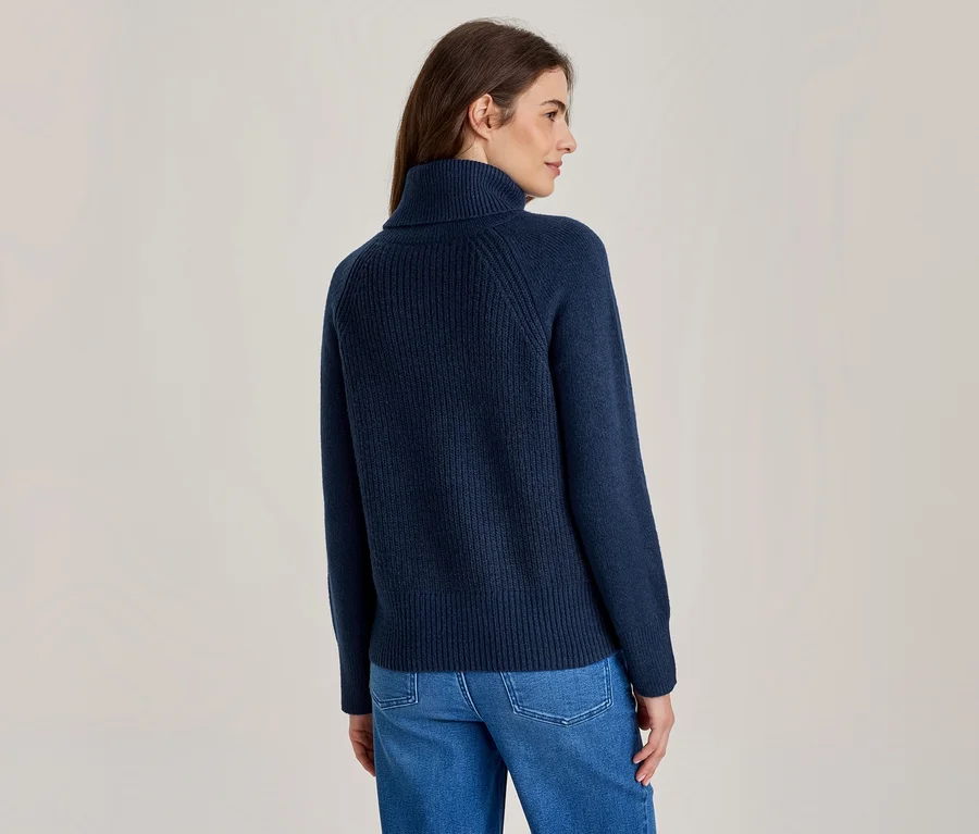 Frau von hinten, leicht gedreht, trägt einen marineblauen gerippten Rollkragenpullover und blaue Jeans.