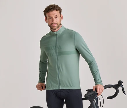 Ein Mann mit kurzen lockigen braunen Haaren und leichtem Bart trägt eine salbeigrüne, langärmelige Radsportjacke mit dezentem Muster und schwarze Radhosen. Er steht und hält den Lenker eines schwarzen Rennrads, während er in die Kamera blickt.