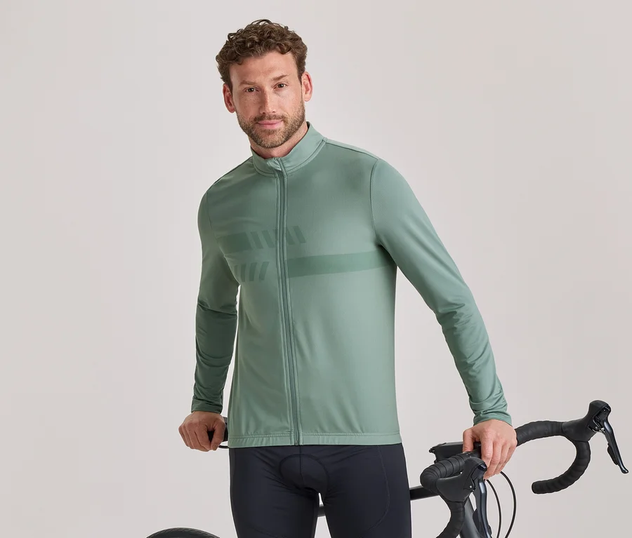 Ein Mann mit kurzen lockigen braunen Haaren und leichtem Bart trägt eine salbeigrüne, langärmelige Radsportjacke mit dezentem Muster und schwarze Radhosen. Er steht und hält den Lenker eines schwarzen Rennrads, während er in die Kamera blickt.