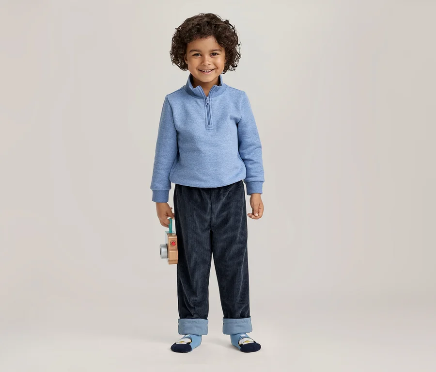 Ein lächelnder kleiner Junge mit lockigem dunklem Haar steht frontal, trägt ein hellblaues Sweatshirt mit halbem Reißverschluss und dunkelblaue Cordhosen mit umgeschlagenen Säumen. Er hält eine hölzerne Spielzeugkamera in seiner linken Hand.